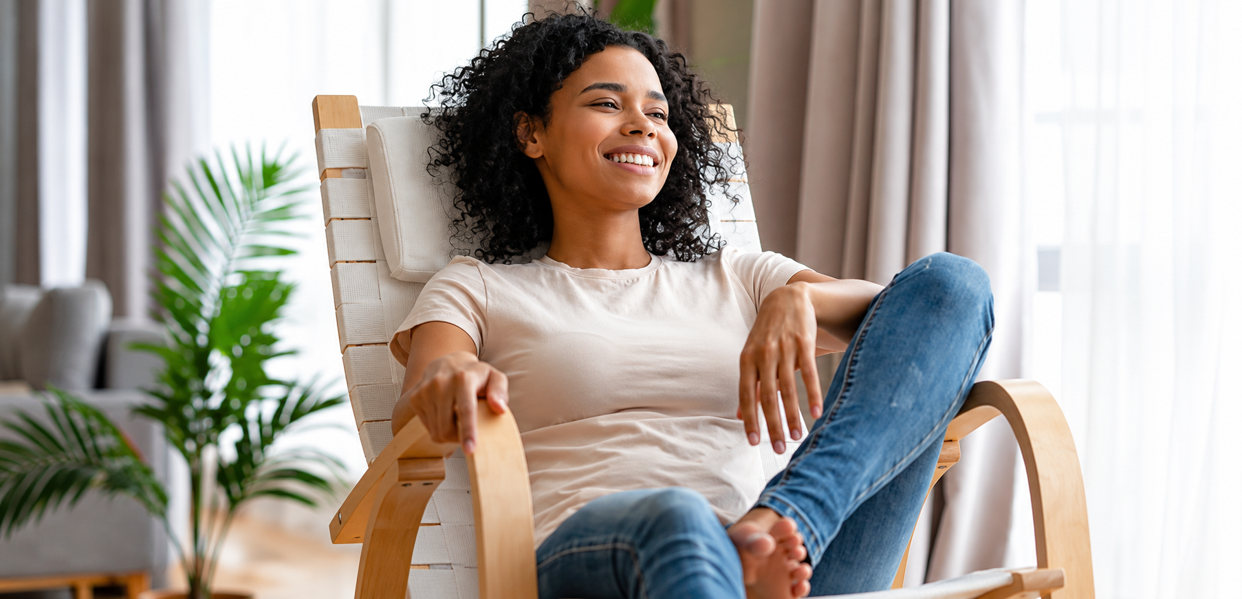 A woman with curly black hair sits smiling in a light wood and white slatted armchair in a bright living room. She is wearing a light beige t-shirt and blue jeans.