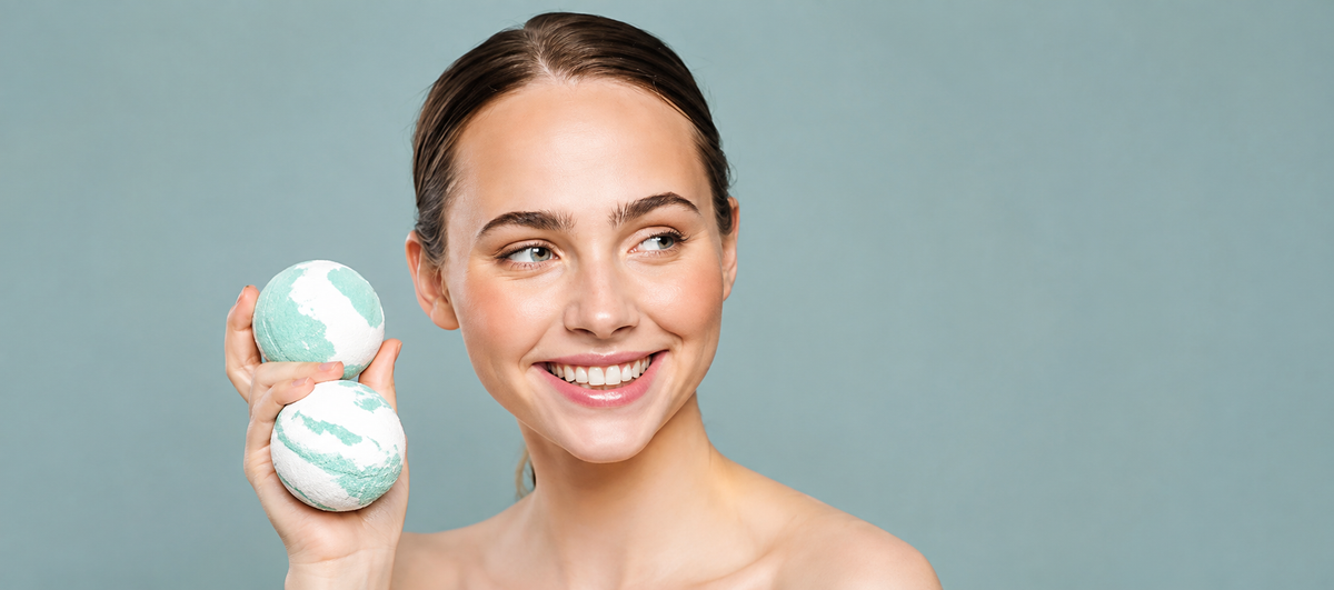 A smiling woman with her hair pulled back holds two round bath bombs with a green and white marble pattern against a solid gray background.