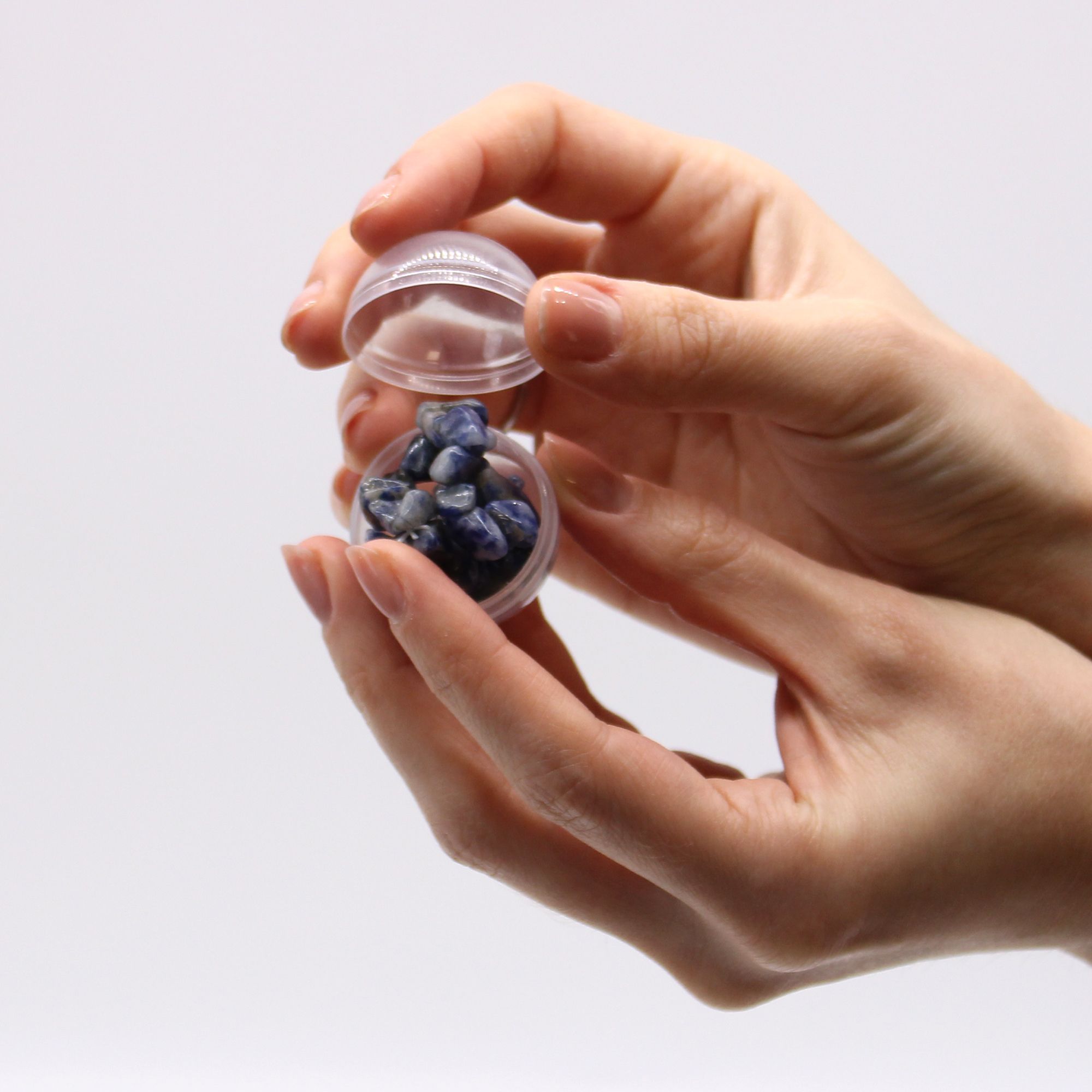 Hand holding a small transparent container filled with a blue bracelet against a light gray background