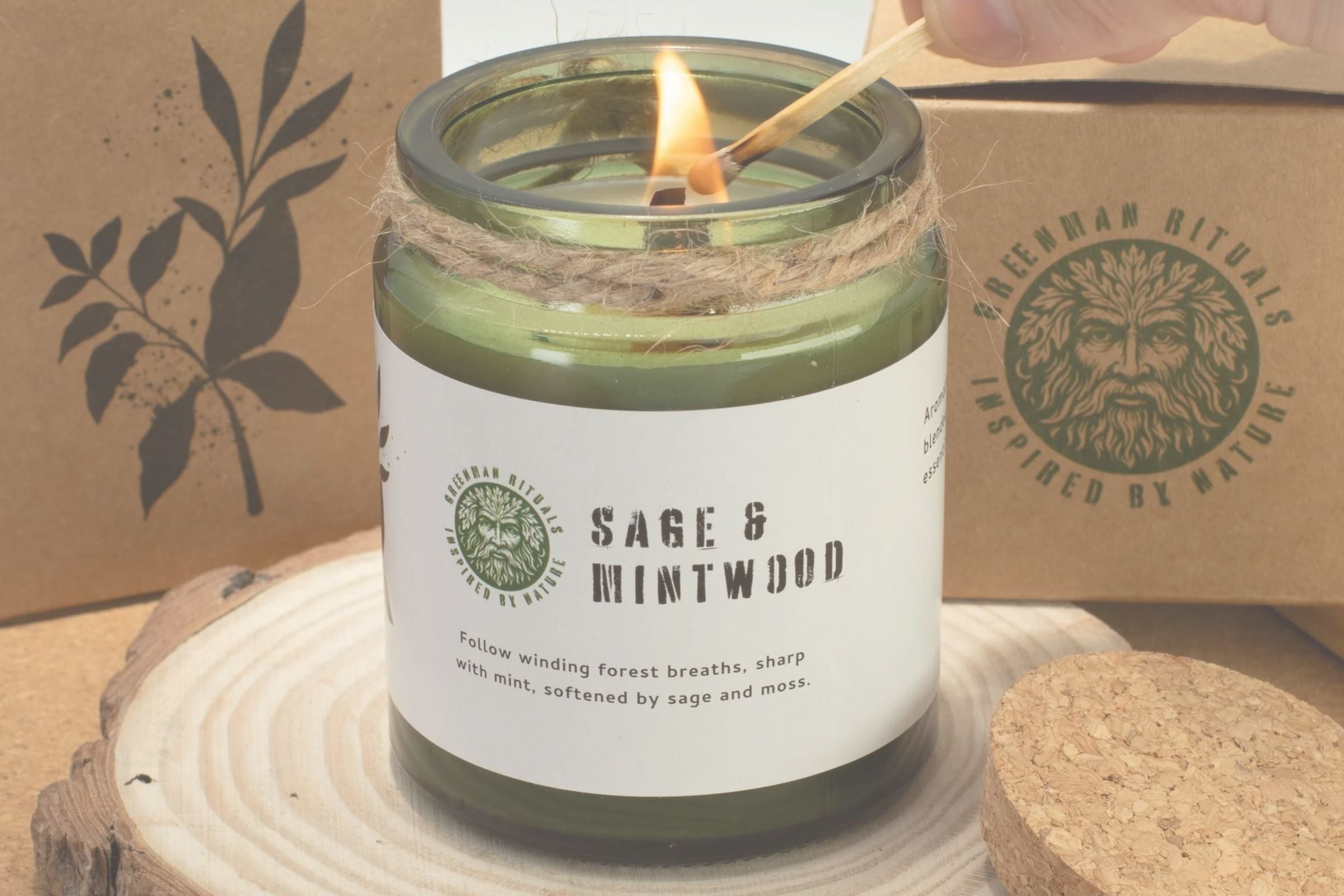 Scented candle labeled 'Sage & Mintwood' being lit with a wooden match, against a brand box background.