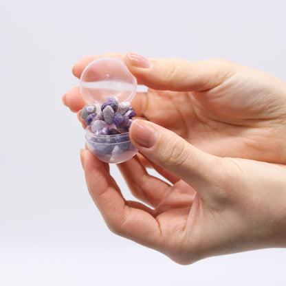Hand holding a small transparent container filled with a purple bracelet against a light gray background