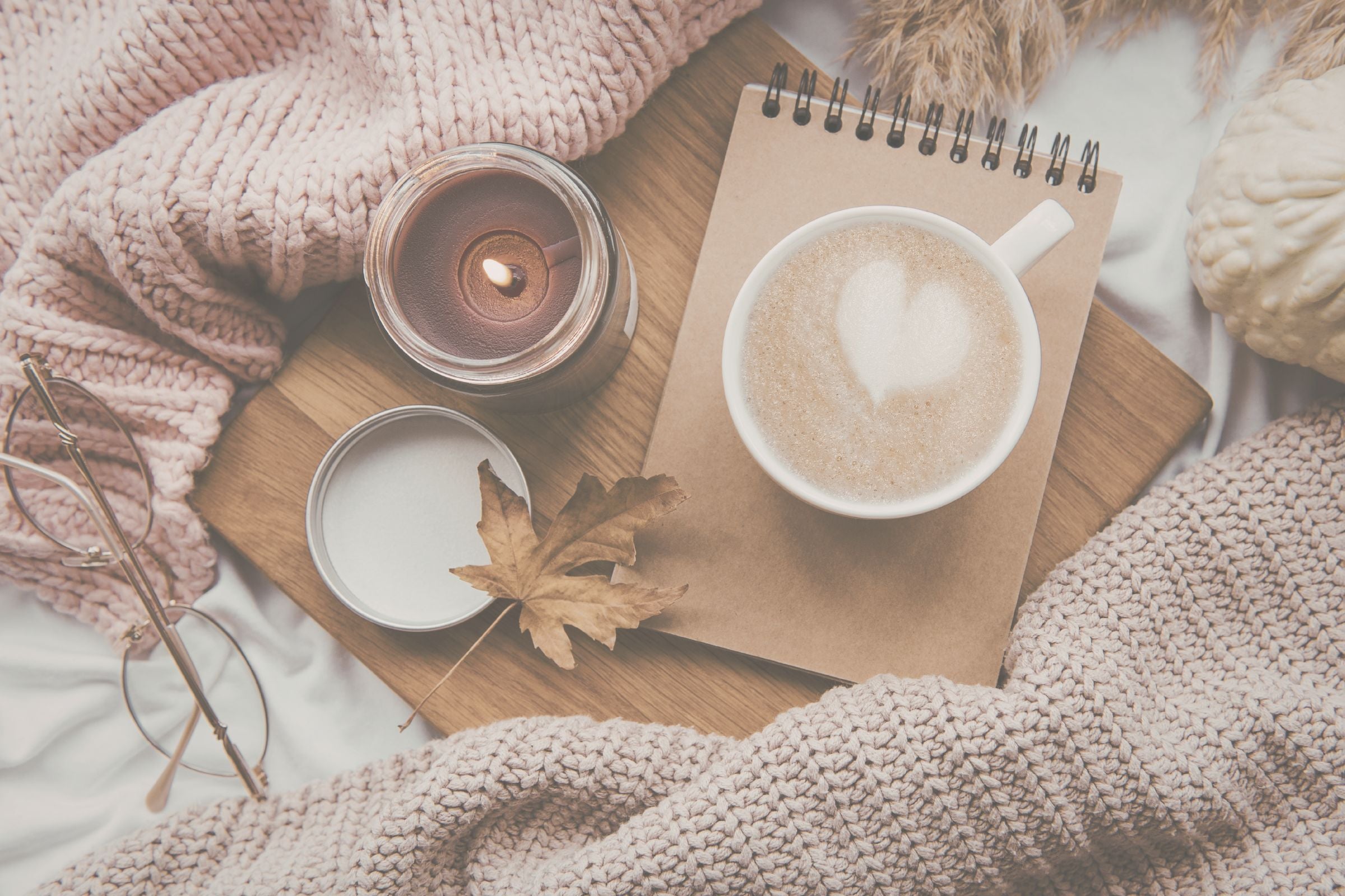 Cup of coffee, notebook, and candle on a wooden surface with cozy blanket and autumn leaves.