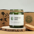 Green candle in a glass jar with a cork lid on a wooden surface, surrounded by nature-themed packaging.