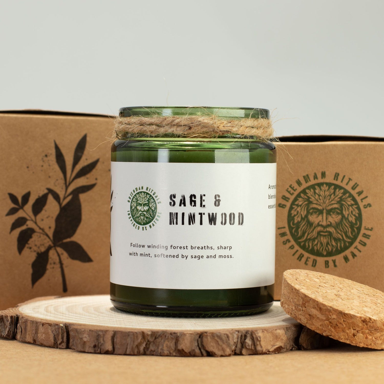 Sage and mintwood candle in a green jar with a label, placed on a wooden surface with a cardboard box in the background.