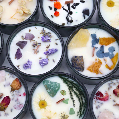 Assorted candles with natural elements like gemstones, flowers and leaves in dark green containers.