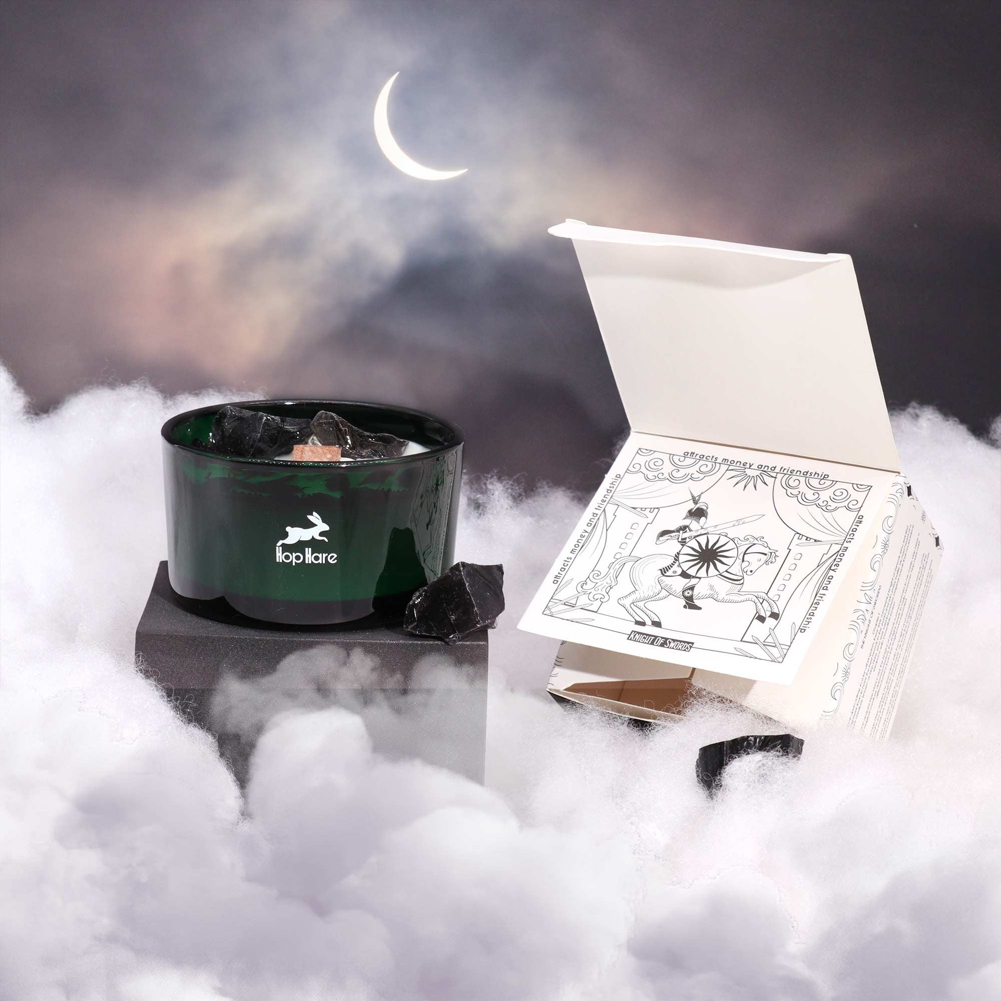 A candle with a green container and a tarot card box on a foggy surface with a crescent moon in the background.