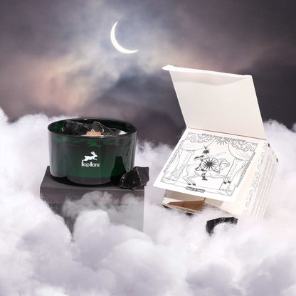 A candle with a green container and a tarot card box on a foggy surface with a crescent moon in the background.
