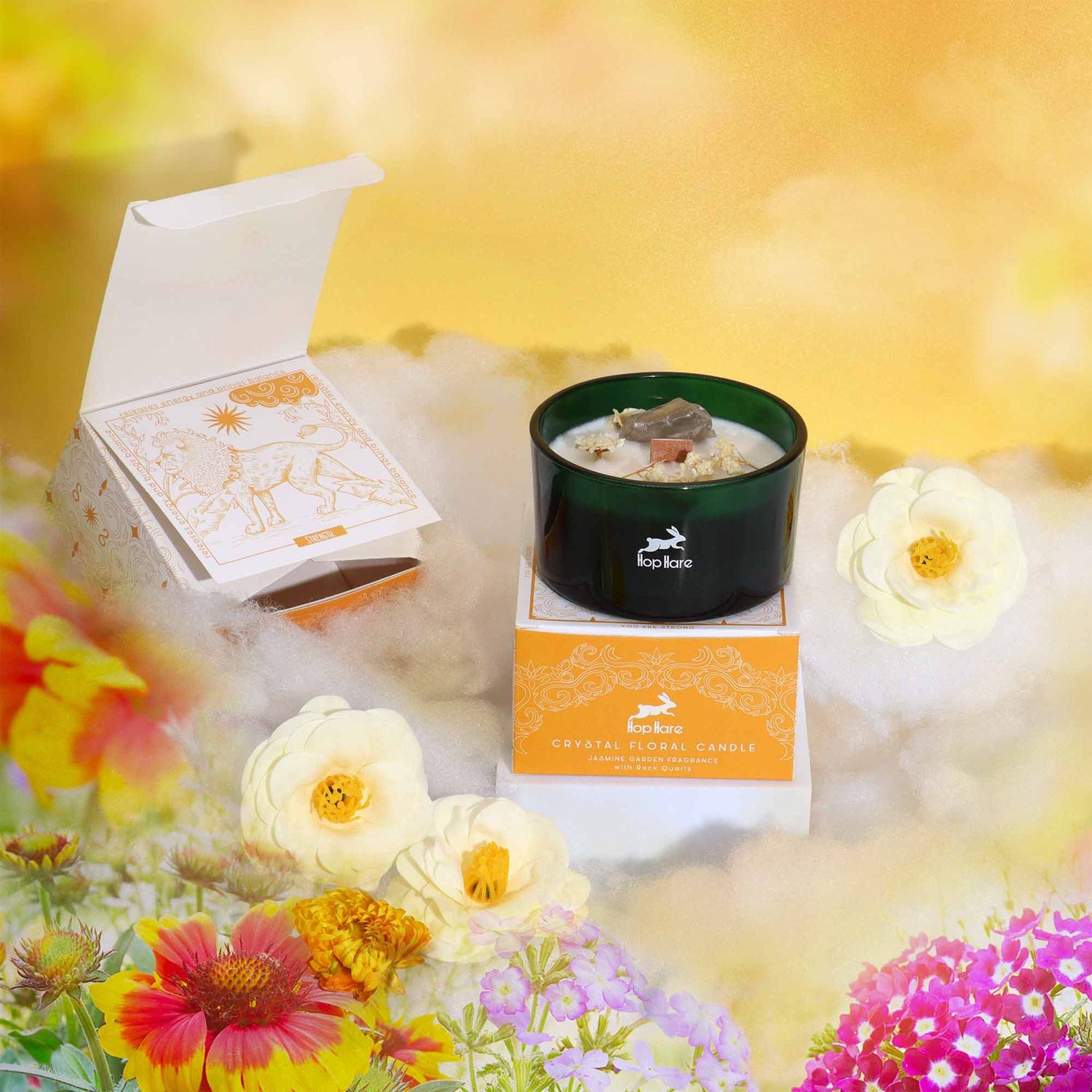 A candle displayed with a tarot card and orange gift box among bright flowers and golden clouds.