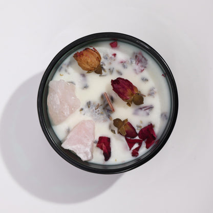 A dark green container with a white candle adorned with rose quartz and dried flowers on a light grey background