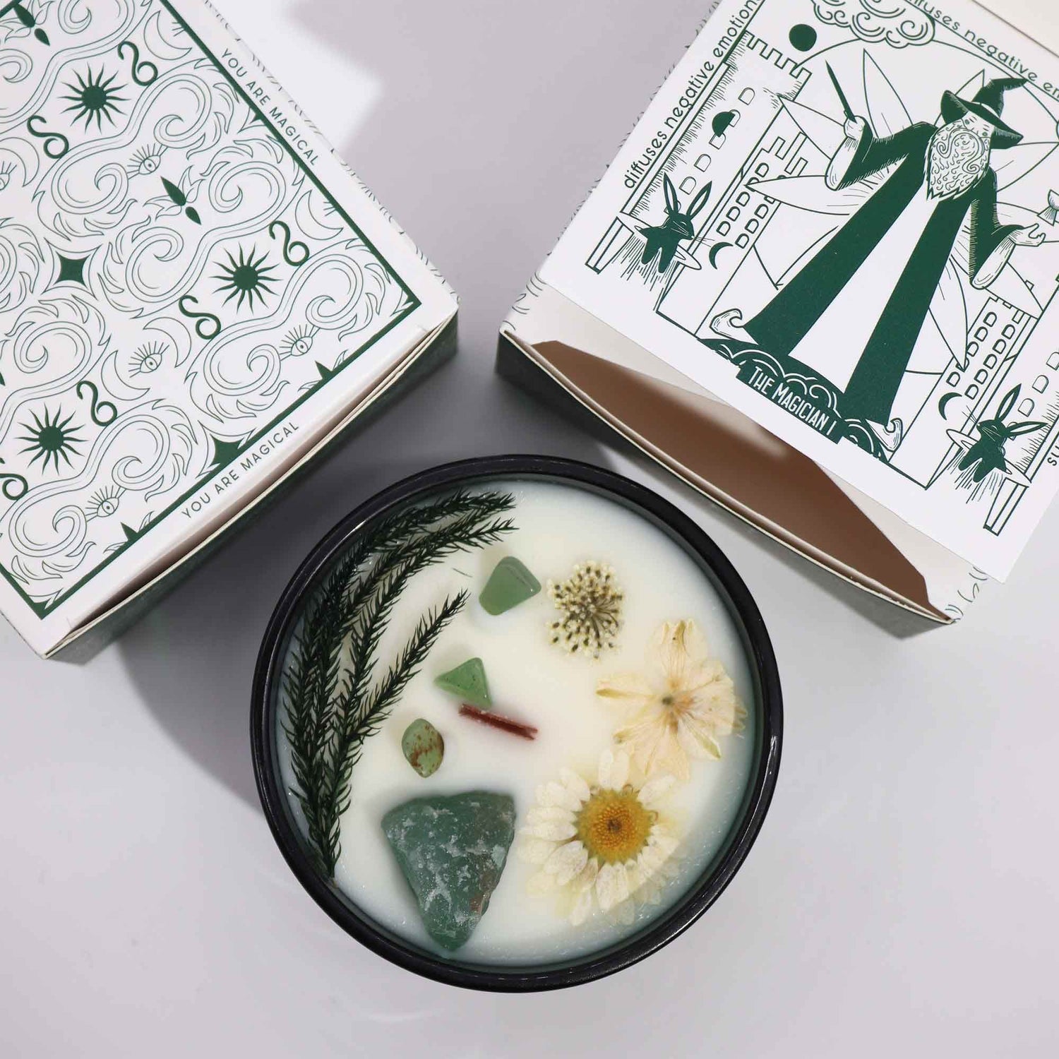 A candle with natural elements in a dark green dish, surrounded by decorative boxes on a light background