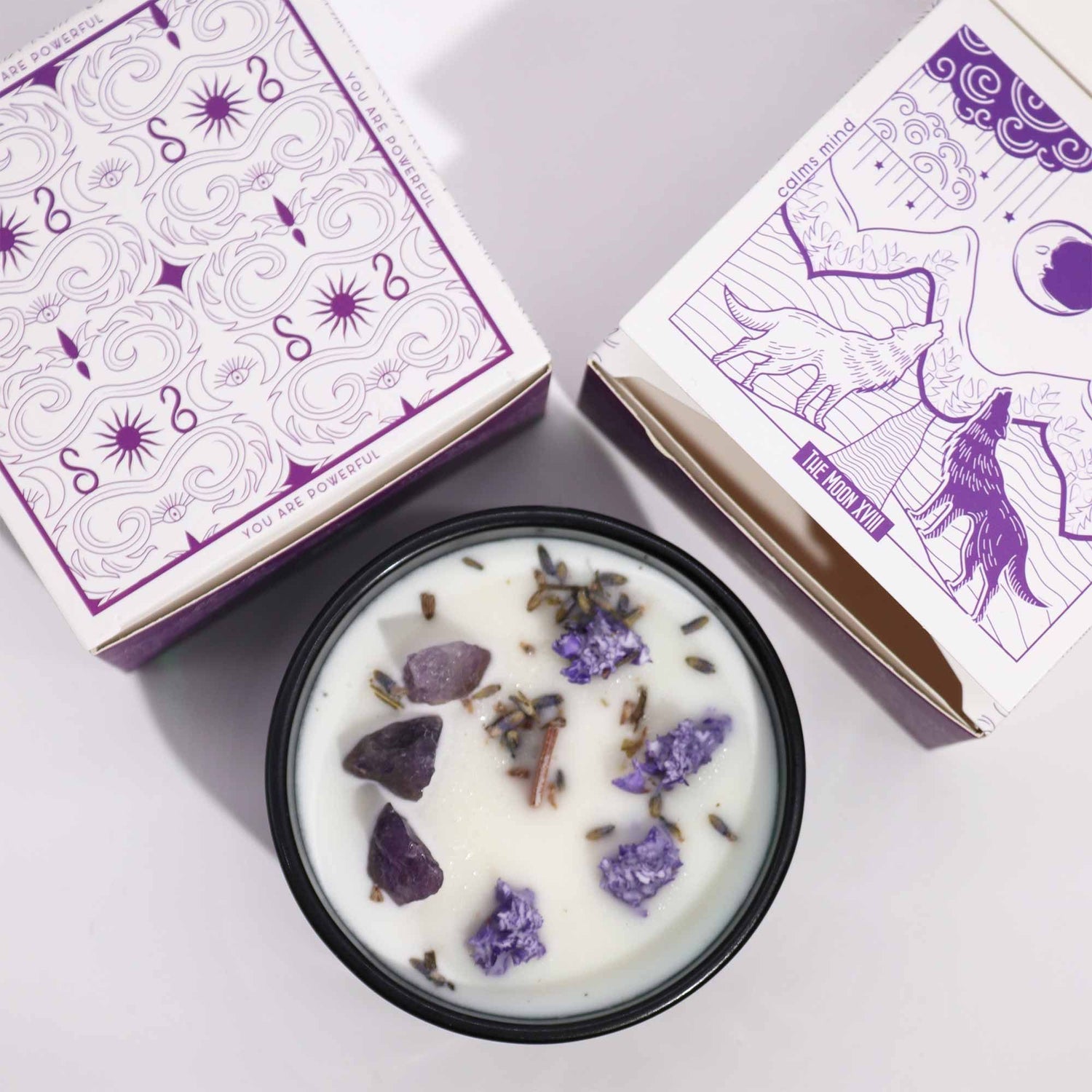 Candle with lavender pieces in a dark green container next to its packaging on a light purple background