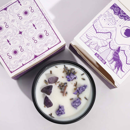 Candle with lavender pieces in a dark green container next to its packaging on a light purple background