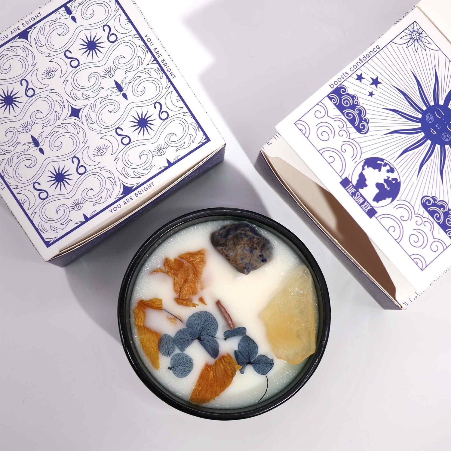 Candle with decorative elements in a black container next to patterned boxes on a light gray background