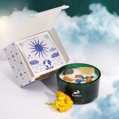 A candle in a dark green container with decorative stones, next to an open box with sun and earth designs on a cloudy background.