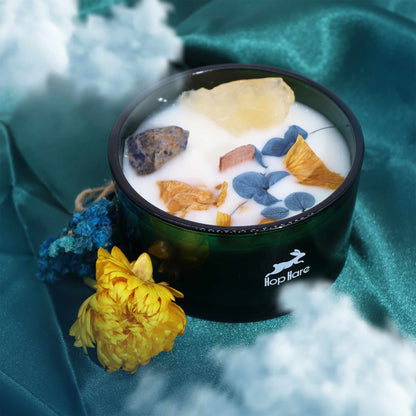 Candle in a dark green container with flowers and rocks on a teal fabric background