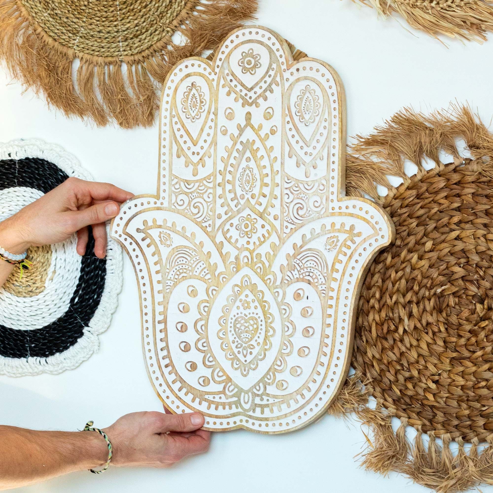 Decorative Hamsa hand wall art with hands holding it, surrounded by woven mats on a white background.