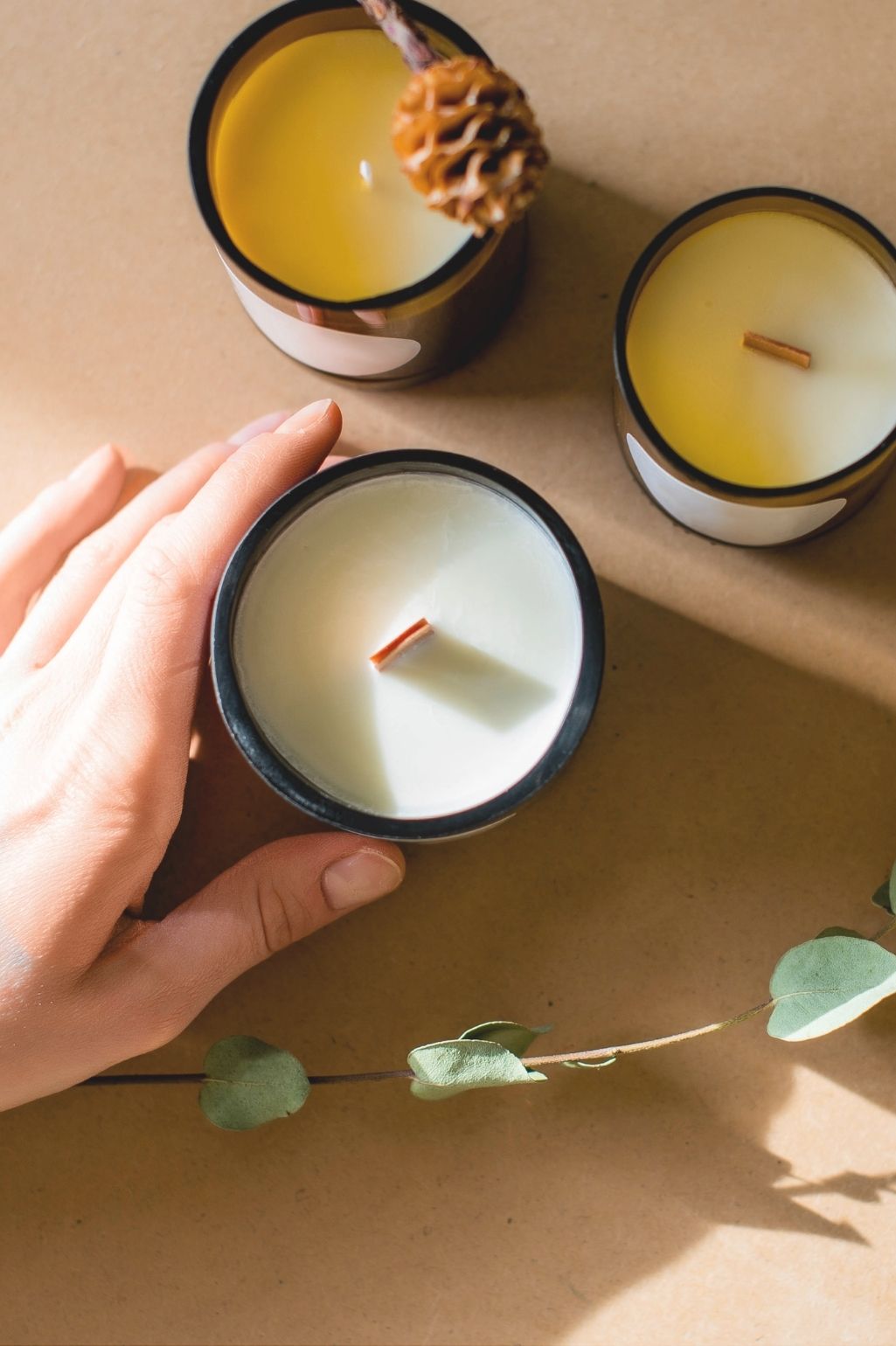 Hand holding a small candle with two other candles on a brown surface.